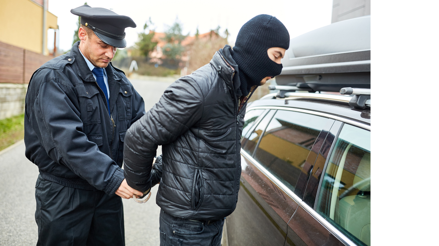 Person in a police uniform handcuffing another person wearing a balaclava next to a car.