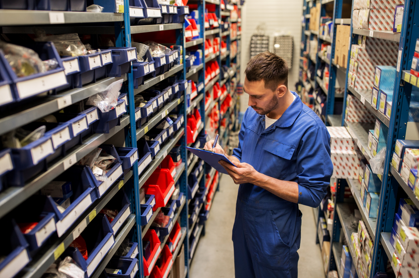 Man in a blue jumpsuit using a tablet in a warehouse setting with shelves stocked with products.